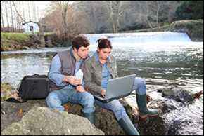 A man and a woman in jeans and fishing vests sit on a rock looking at a laptop with a river in the background behind them.