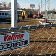 A close up of a metal chain link fence with a sign with the red Valtran logo promoting their fence rentals.